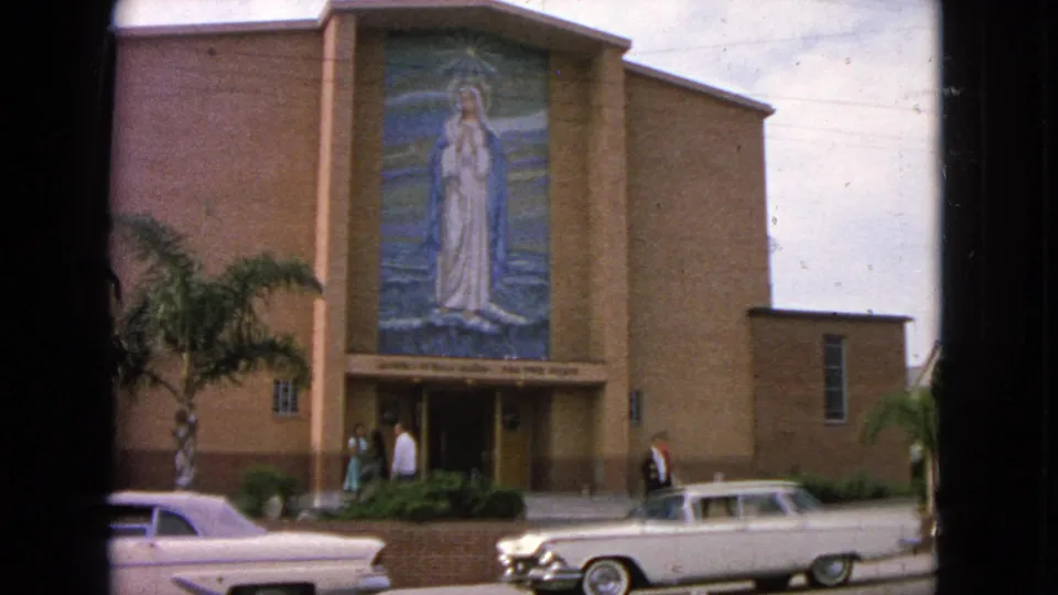 Barrington, Illinois church exterior from 1963 with people near the doorway, a Virgin Mary mural, palm landscaping, and a white early-1960s car at the curb.