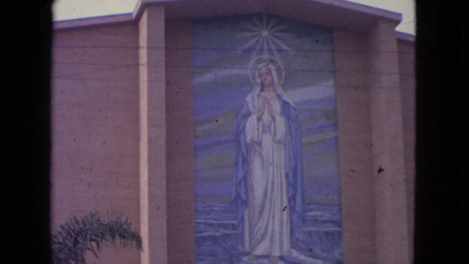 1963 Barrington, Illinois home-movie frame of a large Virgin Mary mural on a church exterior, bordered by dark film gate edges.