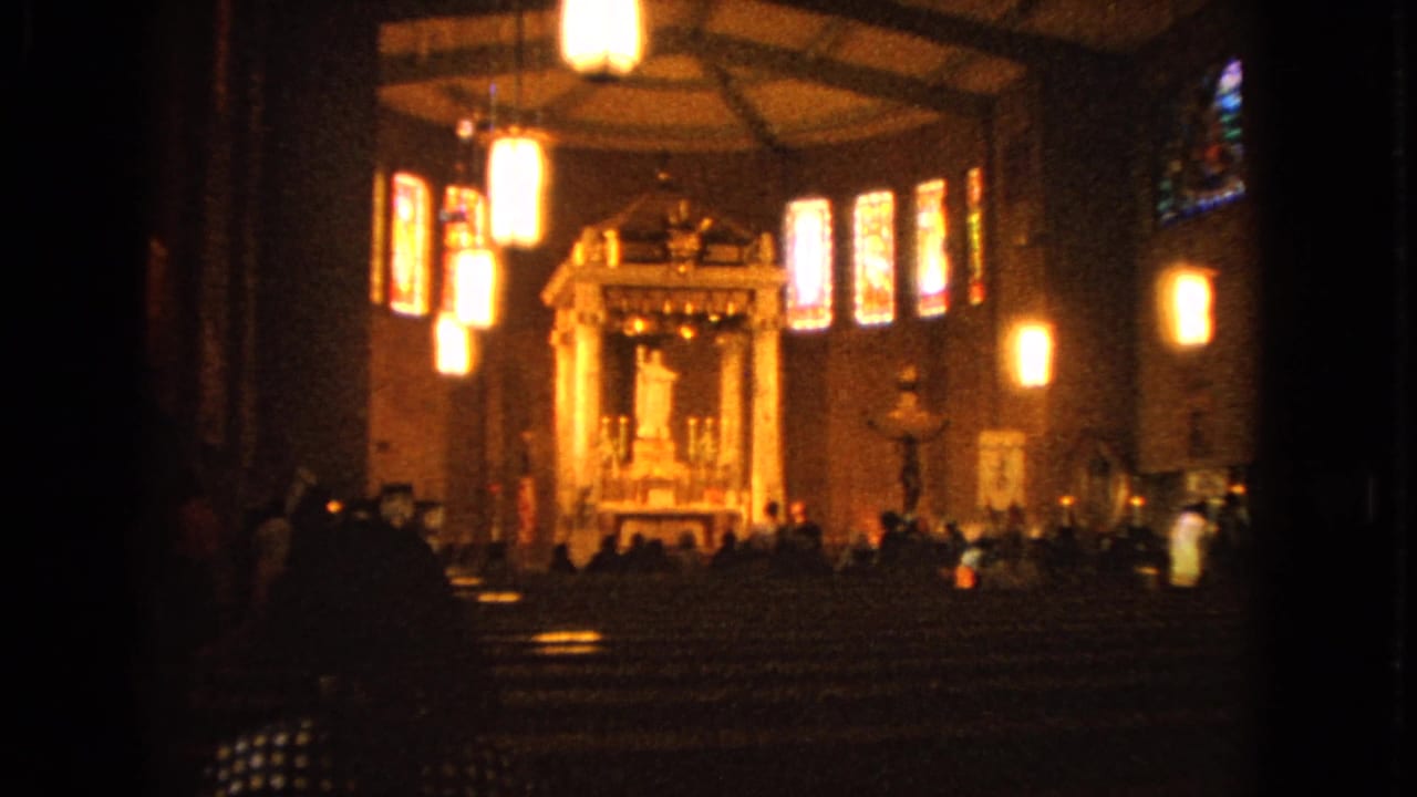 Wide 8mm frame of a Barrington Catholic church interior in 1963, showing a glowing altar area, stained glass, pew rows, and people in the dim foreground.
