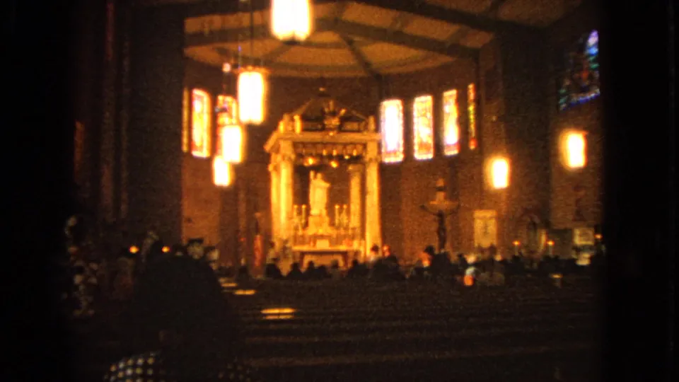 Handheld 1963 8mm view inside a Barrington Catholic church, looking past pews and visitors toward the altar and bright hanging lights.