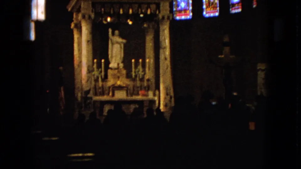 Wide 1963 Barrington church interior with candlelit saint statue, stained glass, and silhouetted worshippers in the pews.