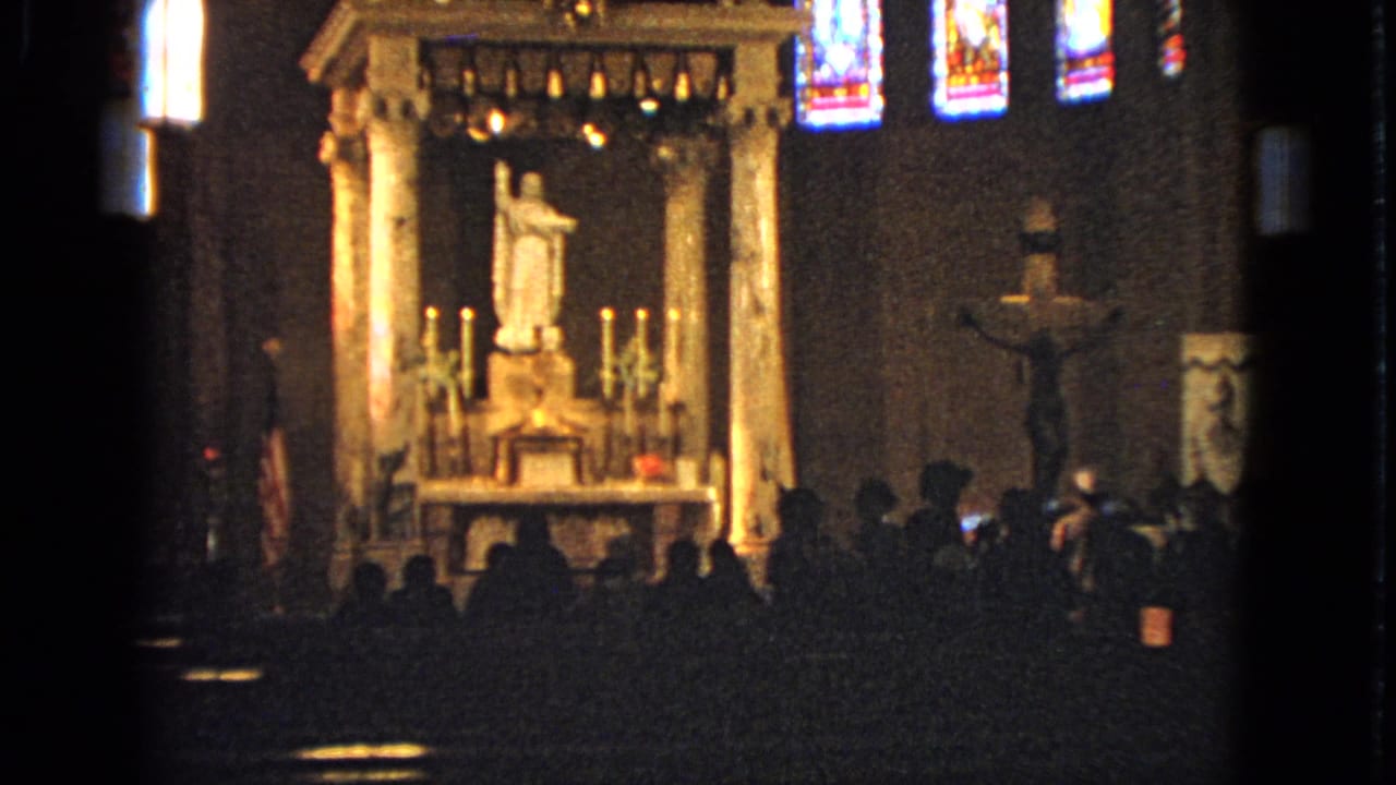 Dim church interior with an illuminated altar, statue, candles, crucifix, stained glass, and seated congregants.