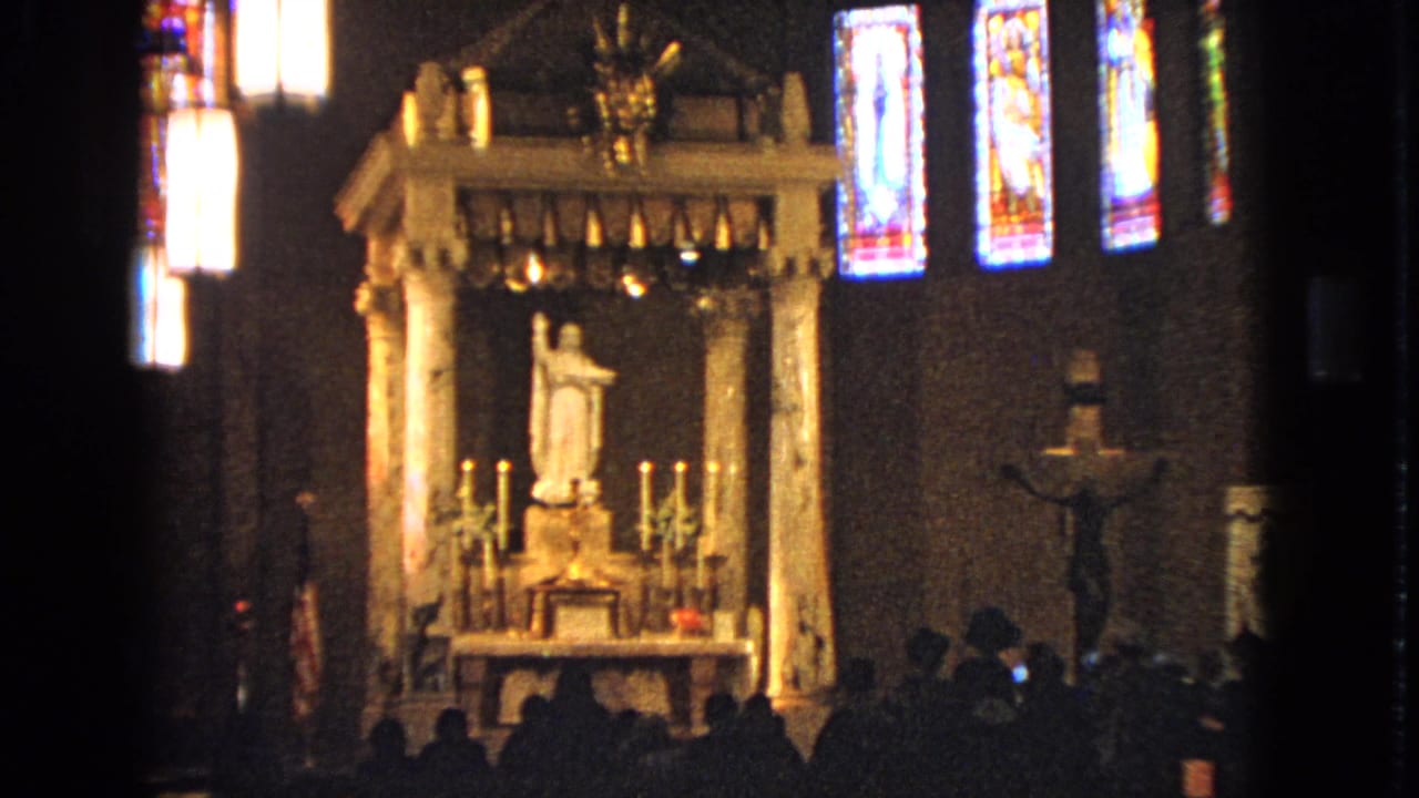 1963 Barrington, Illinois 8mm frame showing a Catholic church altar, statue, candles, stained glass, and worshippers silhouetted in the foreground.