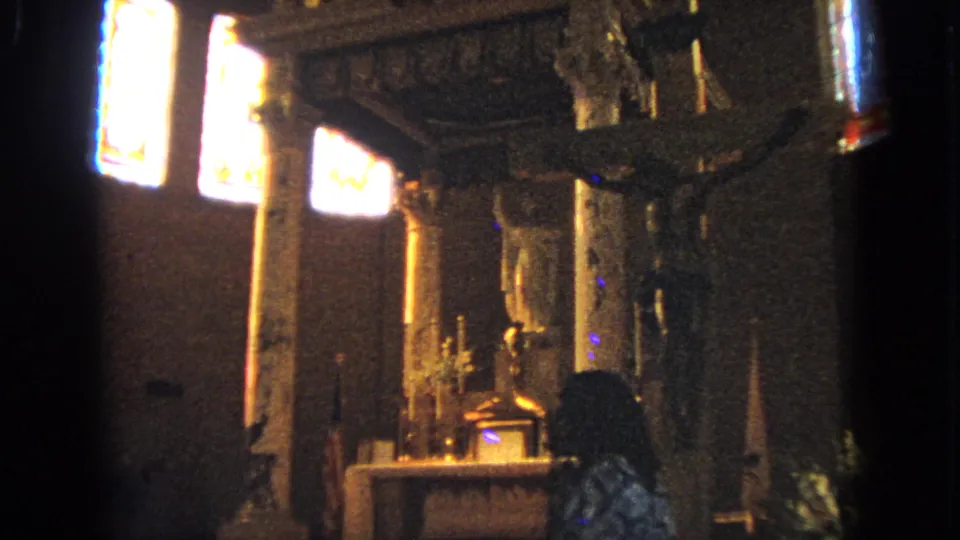 1963 Barrington, Illinois 8mm frame with a closer view of a crucifix near the altar, candles, stained glass, and a dark figure in the foreground.