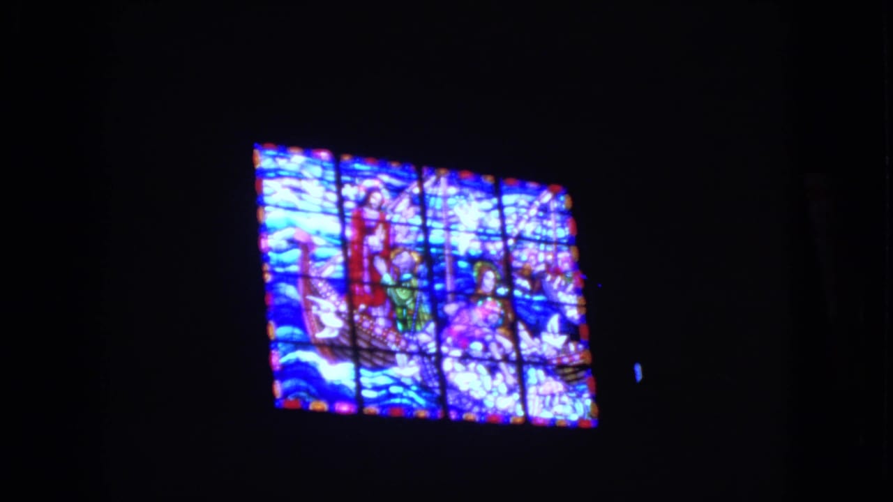 Illuminated stained-glass window in a dark interior, showing a biblical boat scene with blue and red glass.