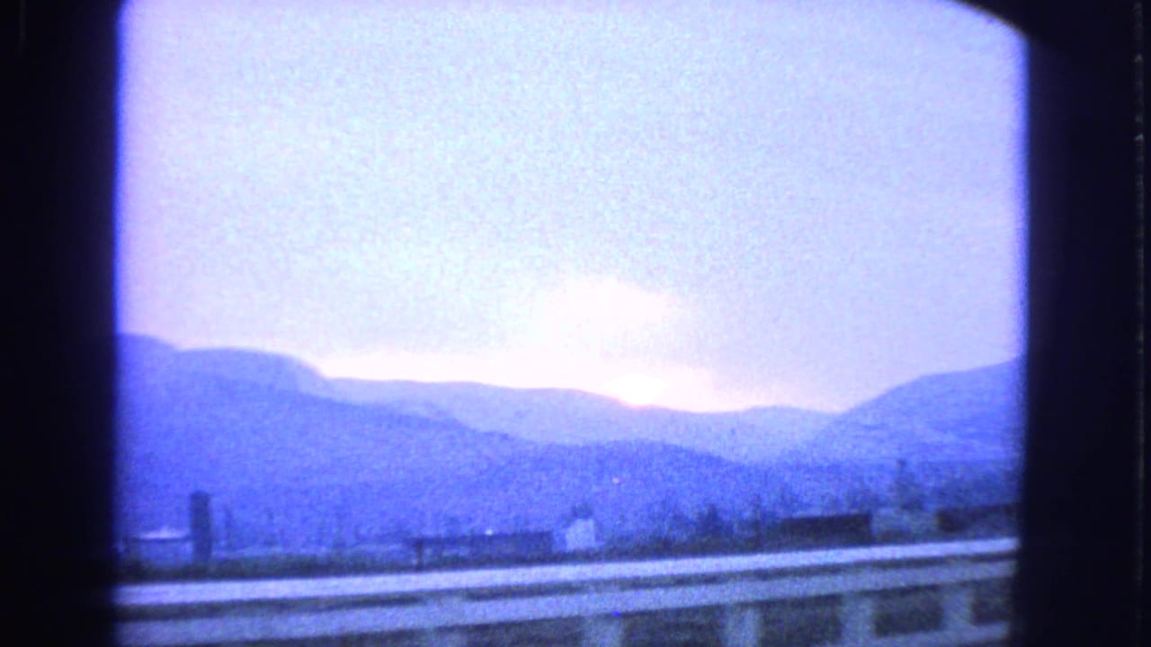 1963 8mm passenger view of distant mountains, low rooftops, and a pale purple dusk sky behind roadside structures.