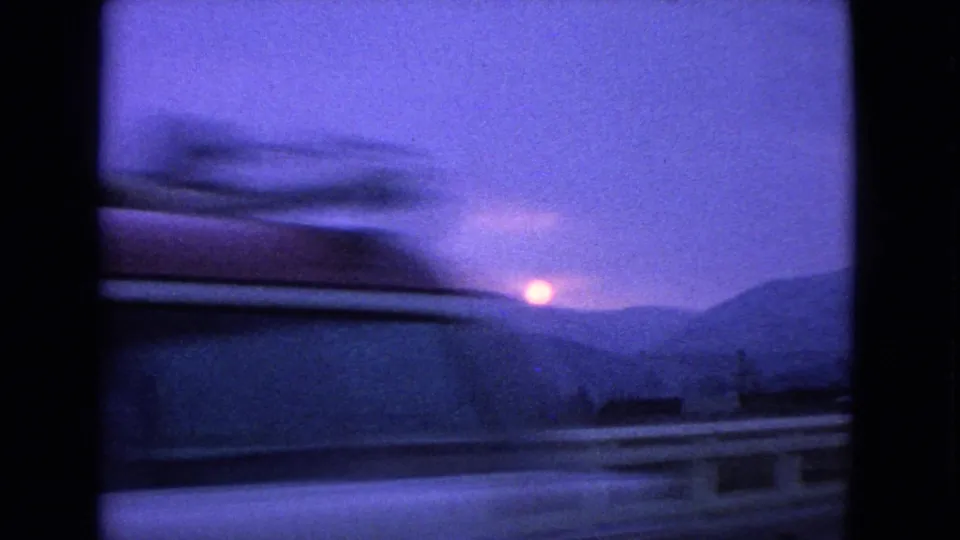 1963 8mm moving-car view with motion-blurred railing, dark mountain ridges, and the low sun near the horizon.