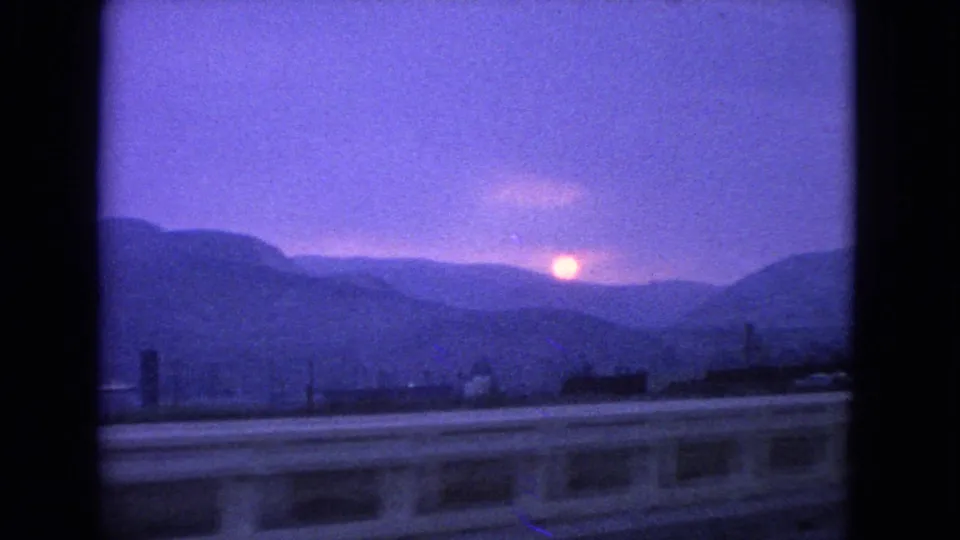 1963 8mm dusk view of the sun over layered mountains, with a roadside railing and small distant buildings in front.