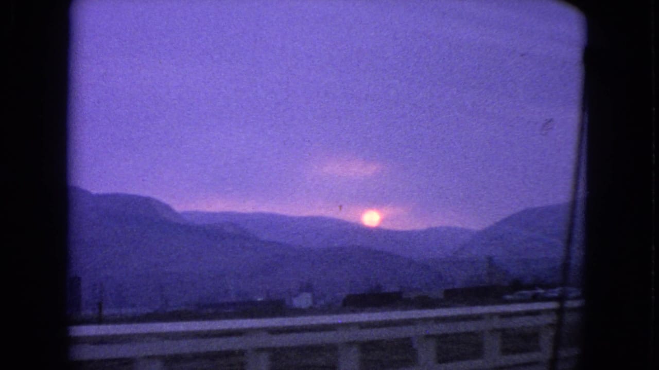 1963 Barrington, Illinois home-movie frame showing a low orange sun beyond distant hills or tree lines, with guardrails and a dark car-window edge in view.