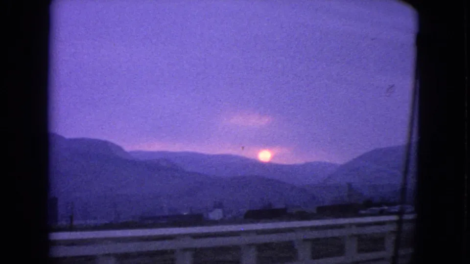 1963 Barrington, Illinois home-movie frame showing a low orange sun beyond distant hills or tree lines, with guardrails and a dark car-window edge in view.
