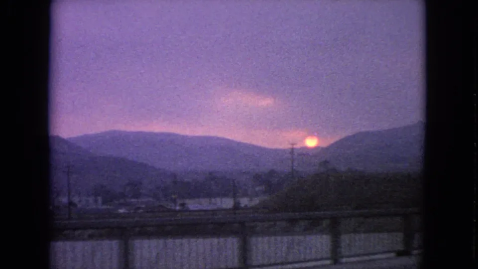 1963 Barrington, Illinois road-view frame with the sun near the horizon, distant ridges or tree lines, utility poles, rooftops, and foreground guardrail.