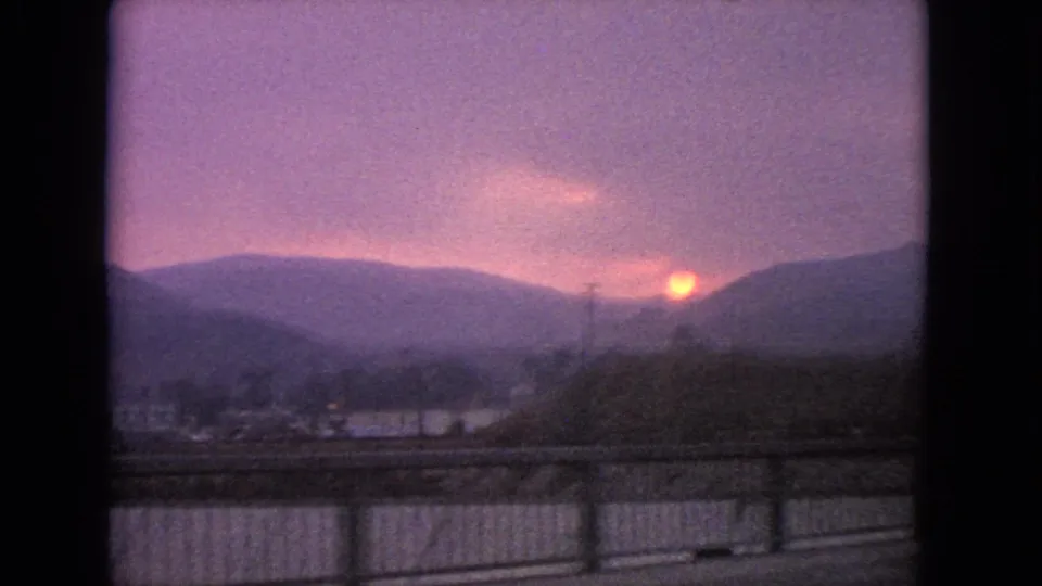 1963 Barrington, Illinois 8mm frame of a soft purple sunset landscape, with the sun near a dark ridge and roadside structures sliding past below.