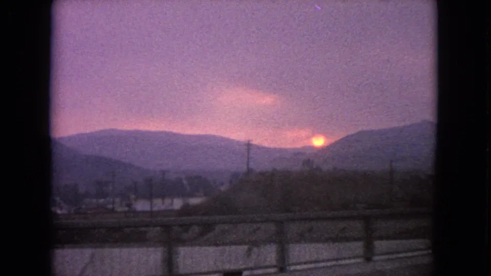 Final 1963 Barrington, Illinois road-shot frame showing the orange sun low over trees or ridges, with guardrails, utility poles, and car-window edges visible.