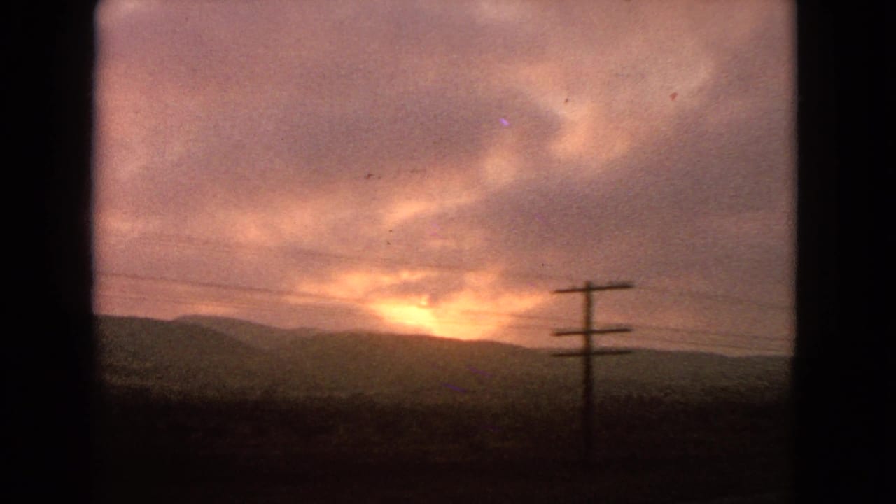 Warm 1963 home-movie view of a sunset behind low hills, seen past utility wires and a pole from a moving car.