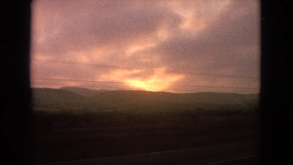 Soft 8mm frame showing a glowing dusk sky over a silhouetted ridgeline, with foreground blur from vehicle movement.