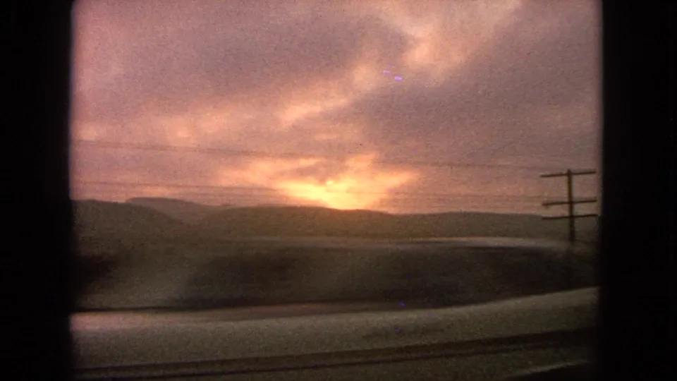 1963 8mm car-window view of a sunset over hills, with utility poles, wires, and a pale roadside or guardrail edge in the foreground.