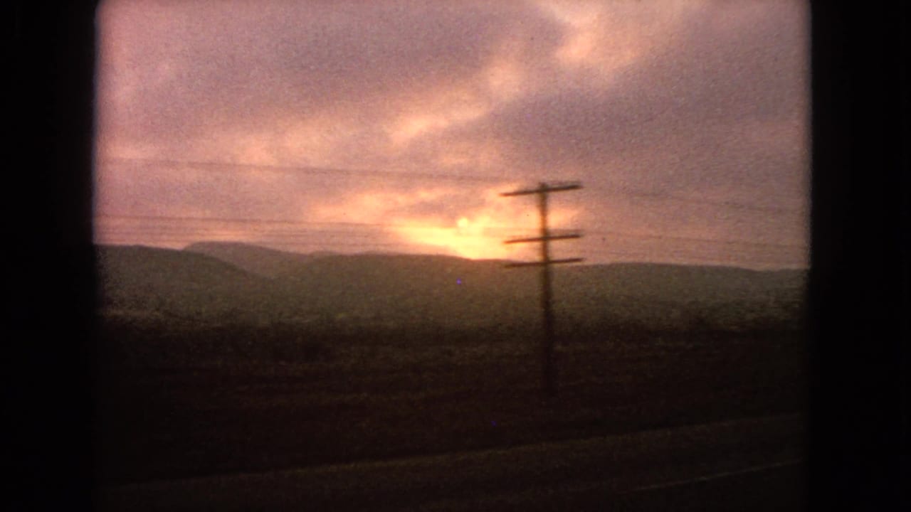 1963 8mm travel footage showing a cloud-covered low sun over dark distant hills, seen past a roadside utility pole.
