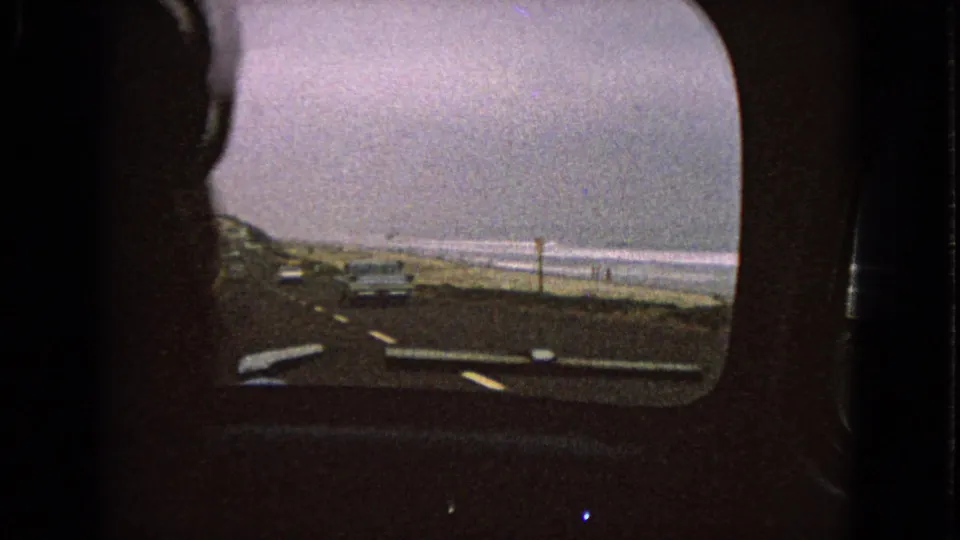 Passenger-side 8mm view from inside a moving car, looking along a coastal road beside beach sand and breaking waves.