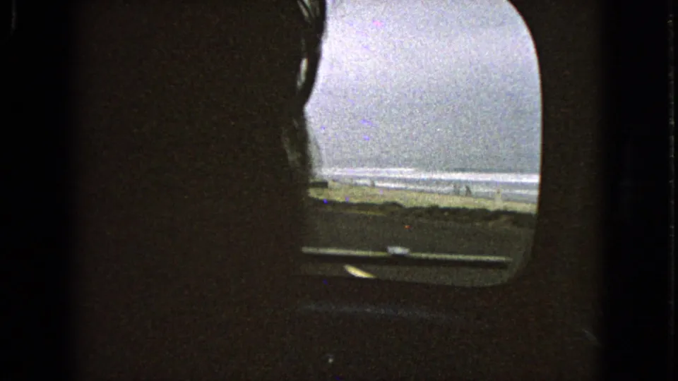 Dim 8mm car-interior frame with a small windshield view of shoreline, surf, and roadway during travel.