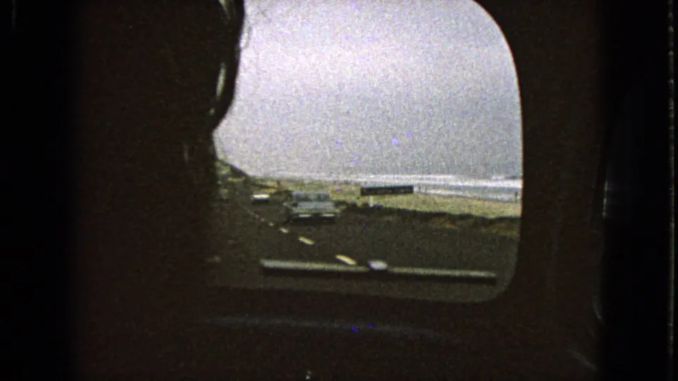 1960s-style 8mm passenger view of automobiles on a coastal road with beach sand and surf visible through the car window.
