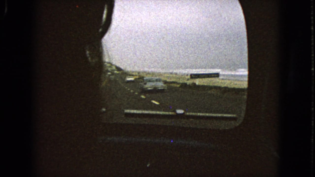 1963 8mm passenger view through a car window toward a roadway beside open water under an overcast sky.