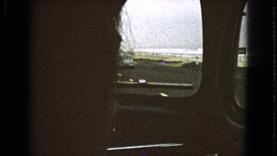 Interior car view in 1963 8mm footage, looking past the window toward a road and water beyond the roadside.