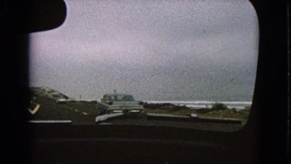 1963 home-movie windshield view of period cars traveling on a road near open water under a gray sky.