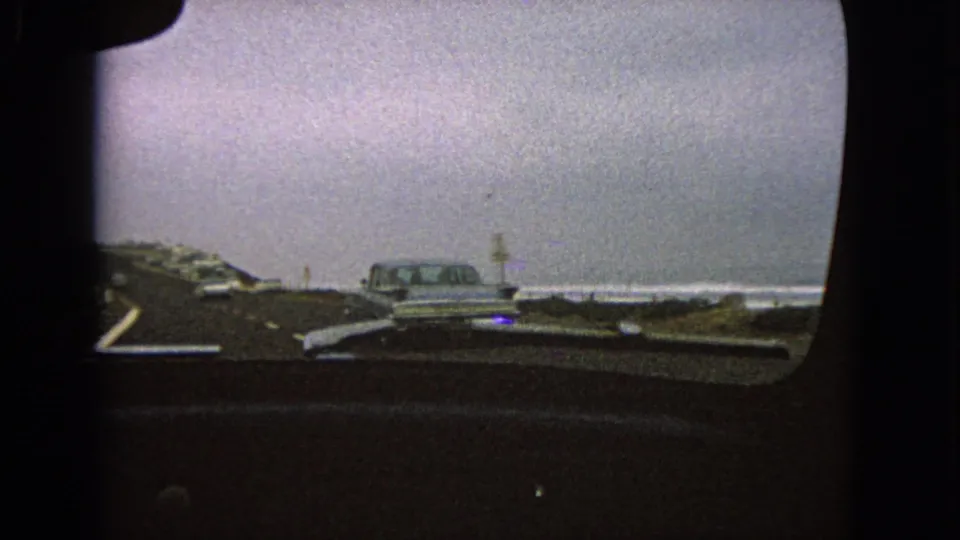 Passenger windshield view from 1963 8mm footage showing a car ahead on a road near open water.