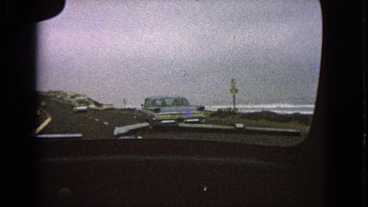 View through a car windshield toward a silver sedan on a curving road near open water.