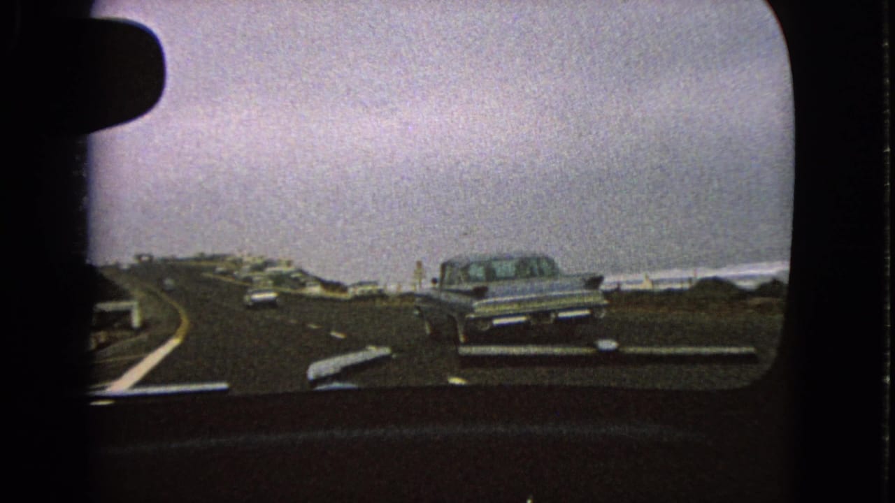 Passenger-side view from a moving car in 1963, with a silver sedan ahead, a curving roadway, and distant water under gray sky.