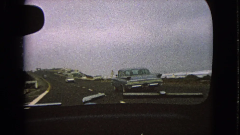 Passenger-side view from a moving car in 1963, with a silver sedan ahead, a curving roadway, and distant water under gray sky.