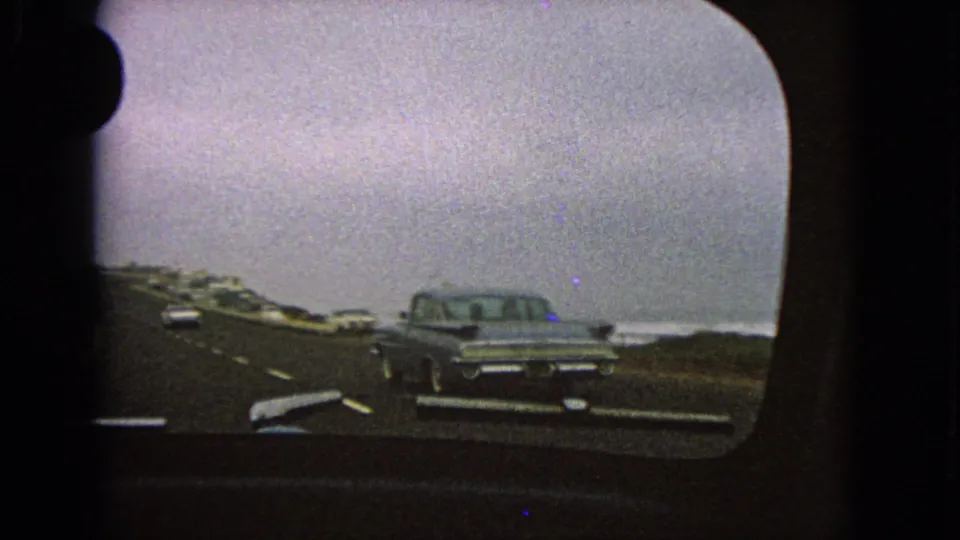 Grainy 8mm frame from inside a moving car, showing a classic sedan ahead on a road with water and low roadside vegetation nearby.