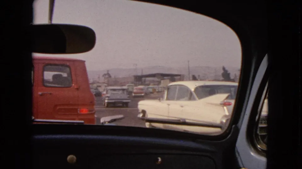 1963 Barrington, Illinois home-movie frame looking through a car rear window at a red van, white sedan, and traffic.