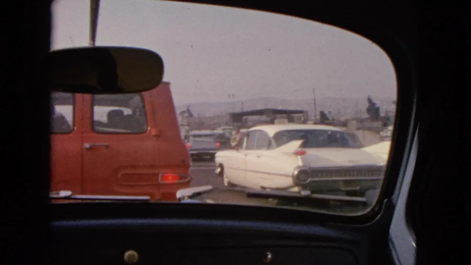 Rear-window 8mm view in Barrington, Illinois, showing a white classic sedan beside a red van on a 1963 roadway.