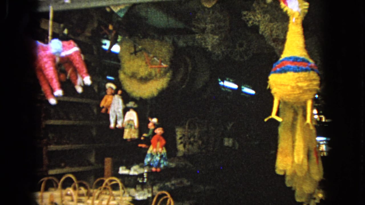 1963 8mm frame showing colorful piñatas, baskets and craft goods hanging or stacked in a dim market display.