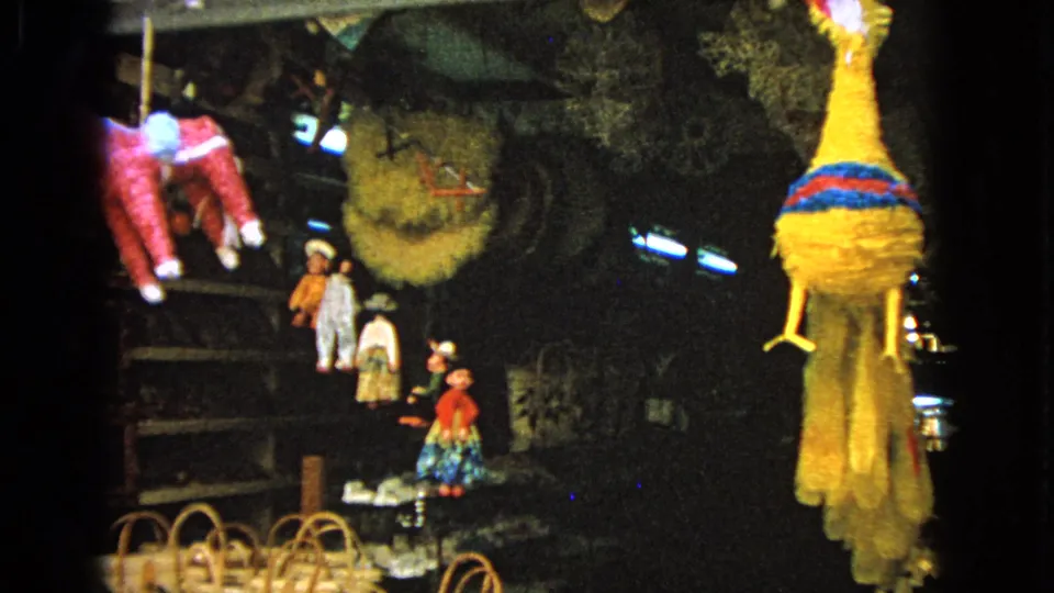 1963 8mm frame showing colorful piñatas, baskets and craft goods hanging or stacked in a dim market display.