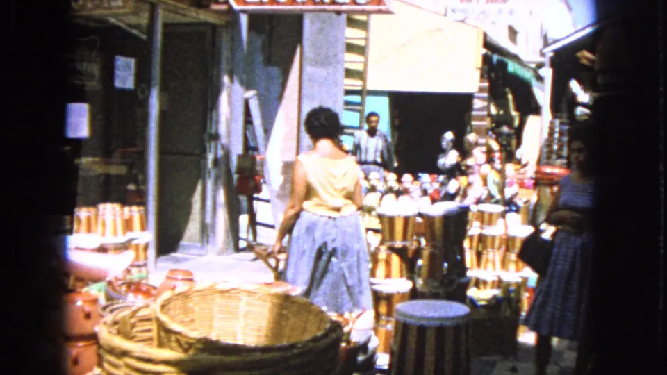 In the 1963 Barrington market footage, a woman in a light top and blue skirt browses baskets, pottery and drums near storefront signs.