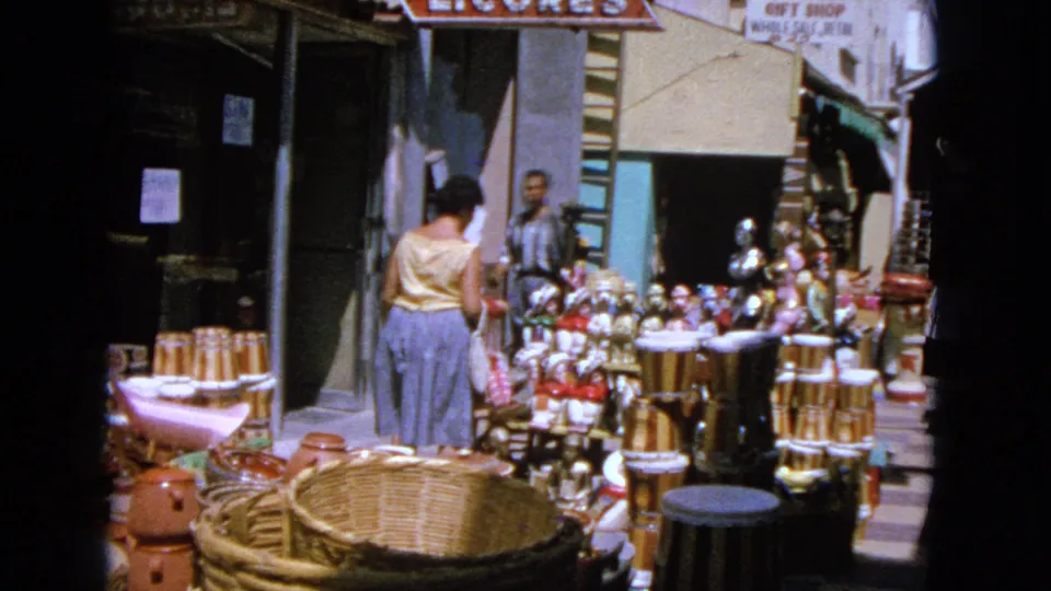 1963 home-movie frame of a woman browsing a dense craft display with baskets, clay pots, drums and figurines in a market setting.