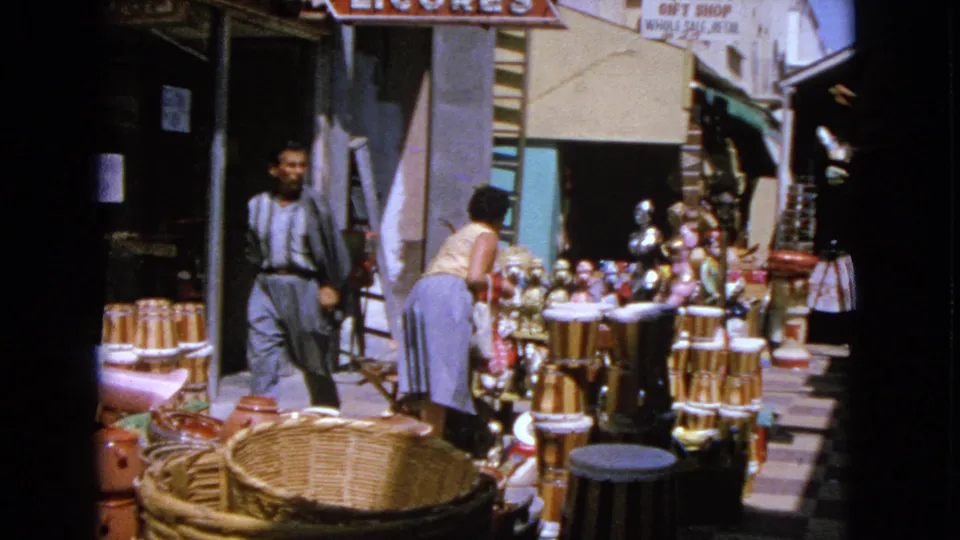 1963 Barrington market frame showing a man walking past as a woman browses baskets, drums, pottery and figurines near a storefront.