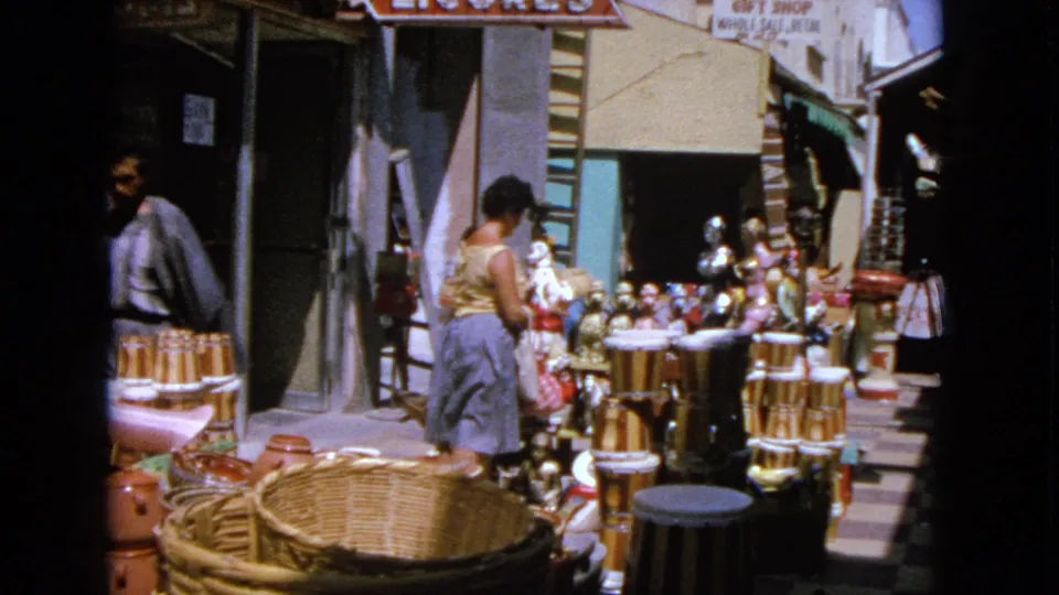 1963 Barrington market scene with a woman in a yellow top and blue skirt looking over baskets, pottery, and figurines near a shop entrance.