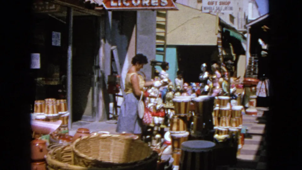 In the 1963 outdoor market setting, the woman studies a dense display of souvenir ceramics, baskets, and small figures beneath storefront signs.