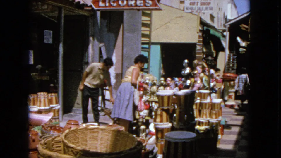 A man in a light shirt appears near the shaded entrance as the woman examines souvenirs at the Barrington outdoor market stall.