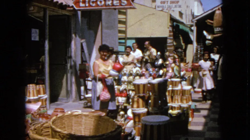 1963 home-movie frame of people browsing outdoor souvenir displays and baskets in Barrington, Illinois, with storefront signs above.