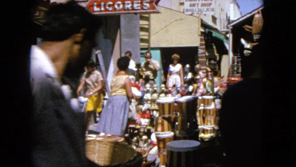 A passerby crosses the foreground while people browse souvenir goods in a sunny 1963 Barrington shopping lane.
