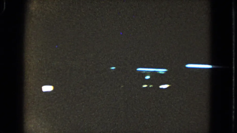 Underexposed 8mm frame showing black gate borders, grain, and scattered bright streaks likely from lights or film exposure artifacts.
