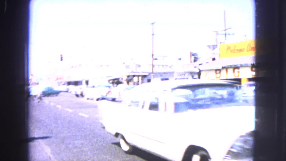 Overexposed Barrington street frame from 1963 with a white classic car, storefronts, traffic lights, and strong daylight glare.