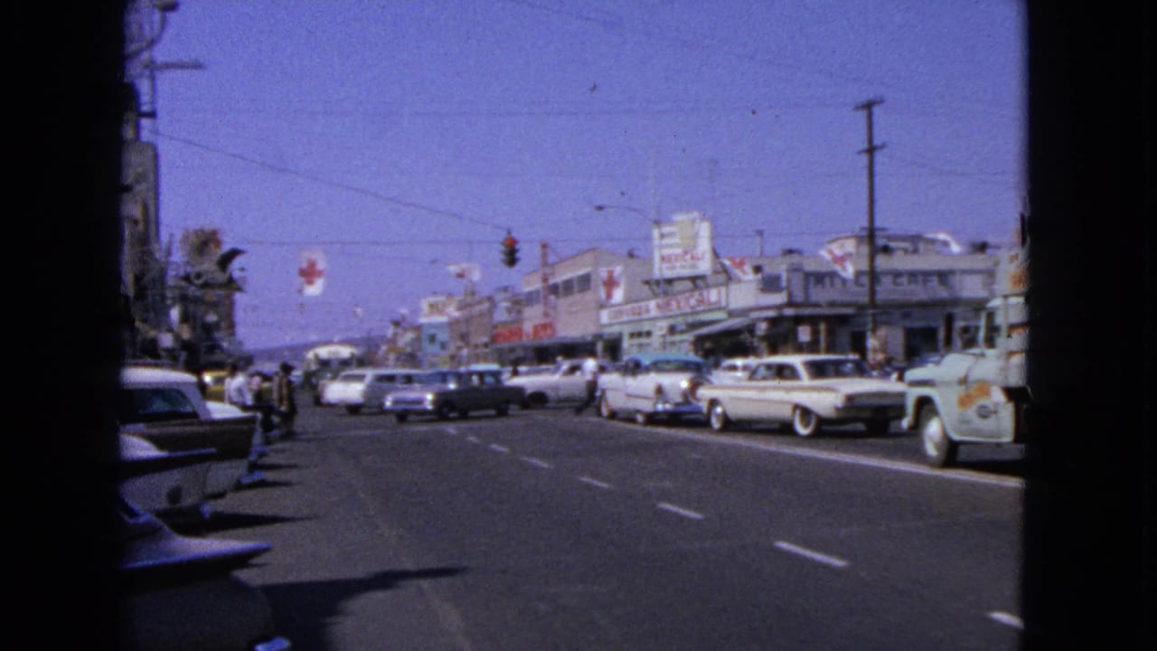 Busy street with parked classic cars, storefronts, traffic lights, and pedestrians in the distance.