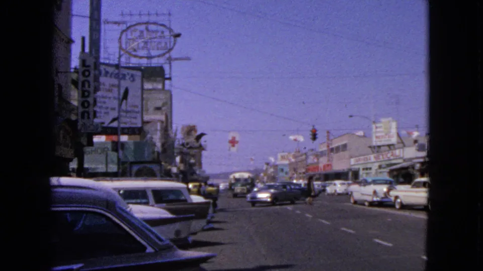 1963 Barrington commercial block with a vertical sign, classic cars at the curb, moving traffic, and pedestrians near the shops.