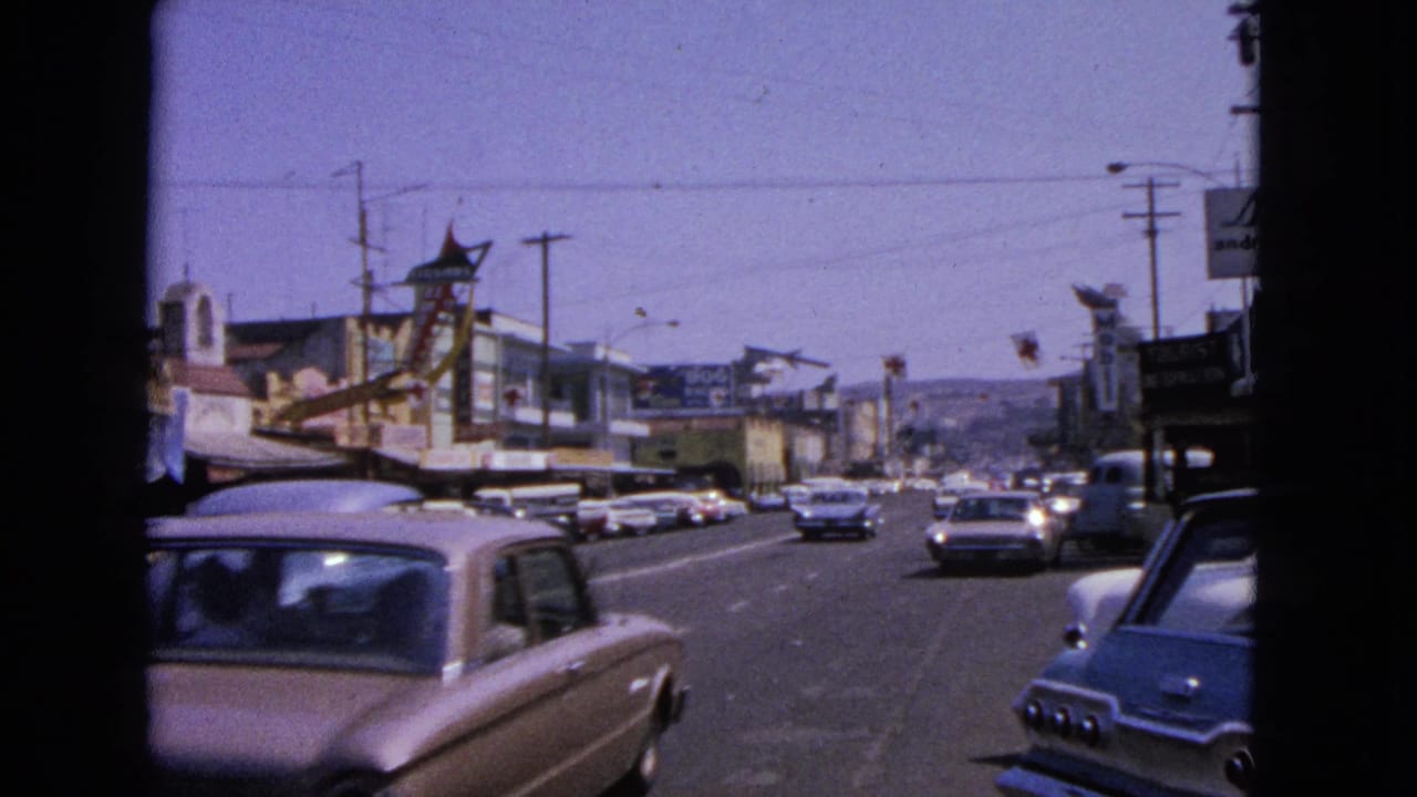 Classic cars drive along a sunny commercial street with storefronts and roadside signs.