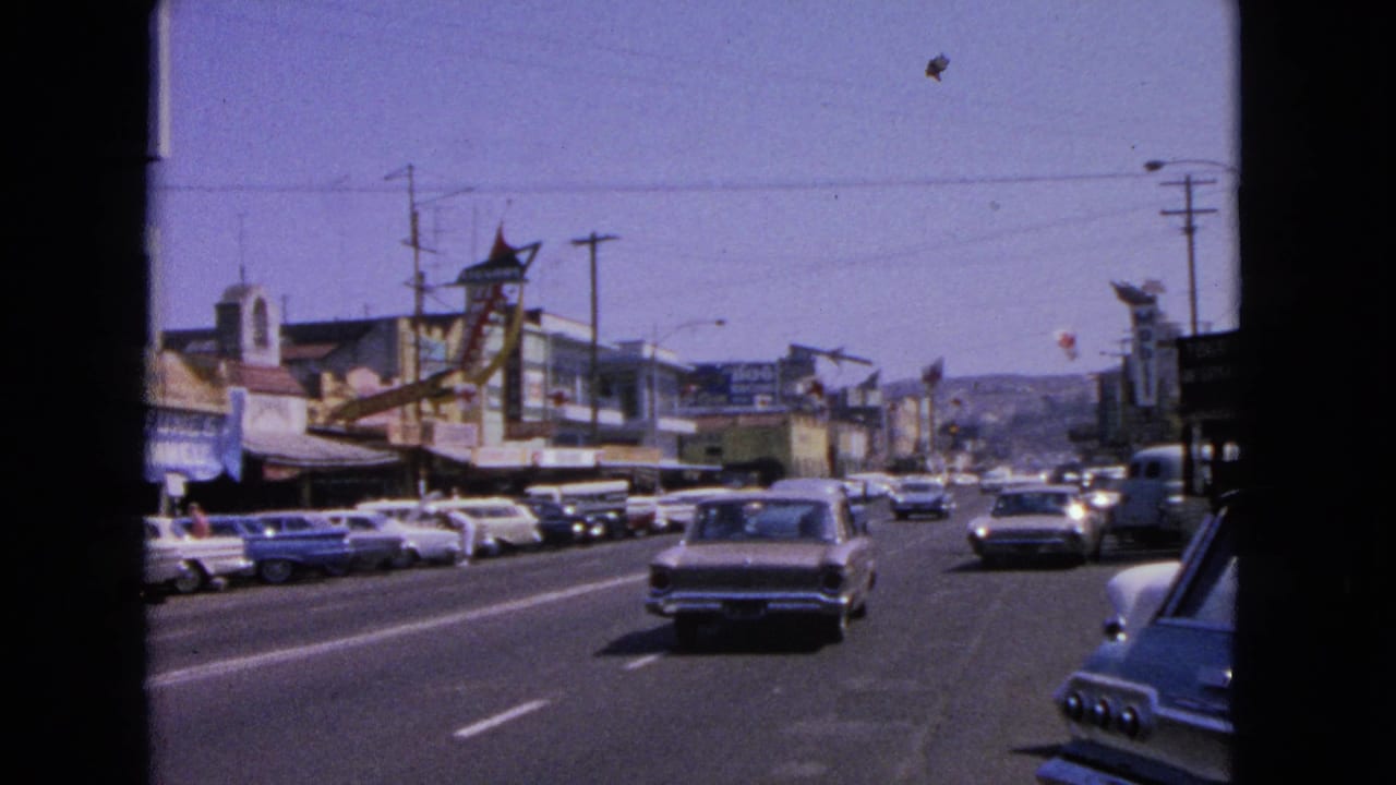 Barrington, Illinois traffic scene showing moving and parked early-1960s cars, storefront signage, utility poles, and a sunlit roadway.